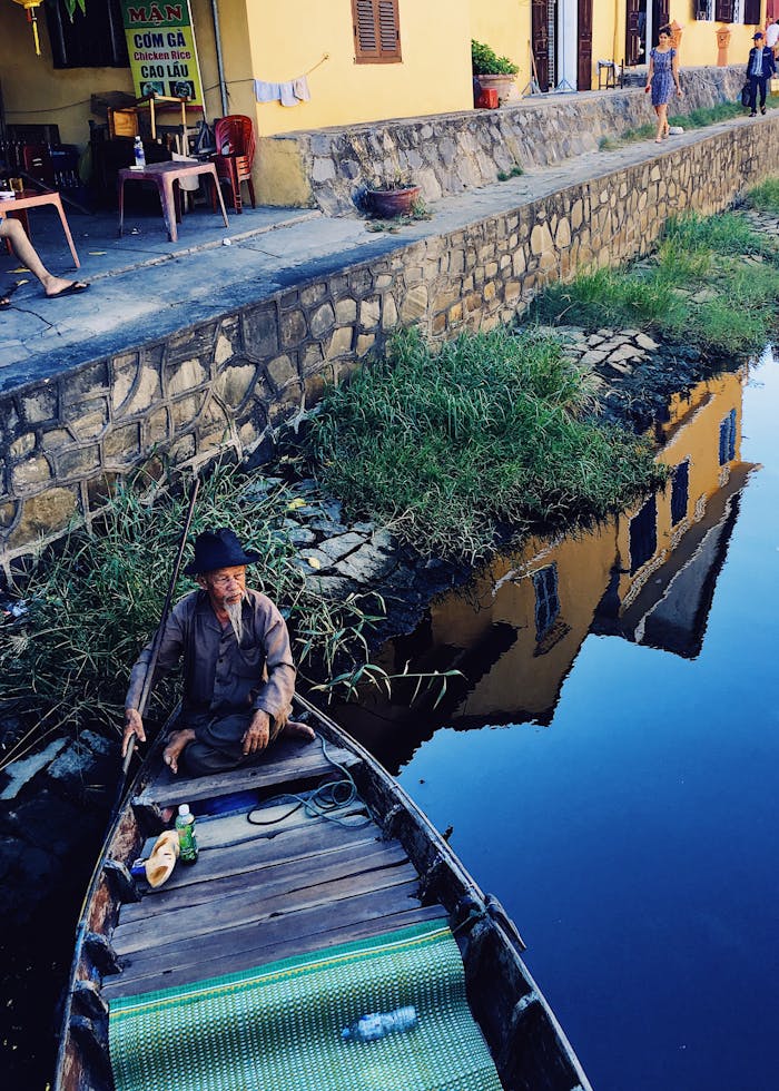 Elderly man navigates a wooden boat along Hội An canal, reflecting the vibrant riverside architecture.